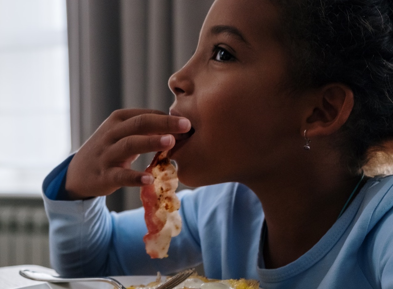 Young girl is eating grilled bacon, looking happy.