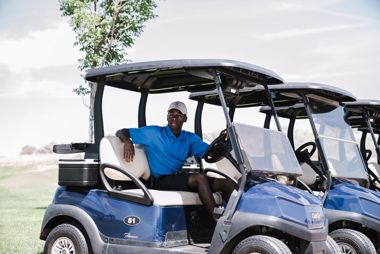 Black man wearing blue shirt and black hat is seating in golf cart.