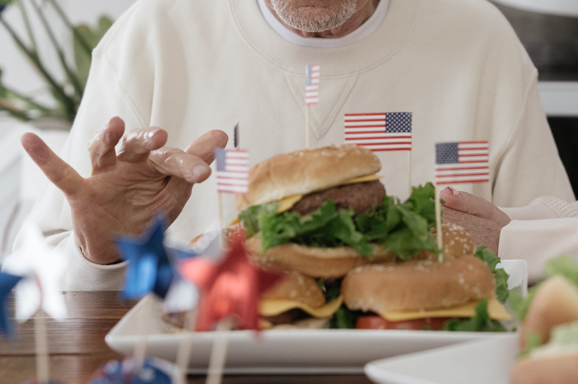 Man wearing white hoodie is siting on the table and eating a burger.