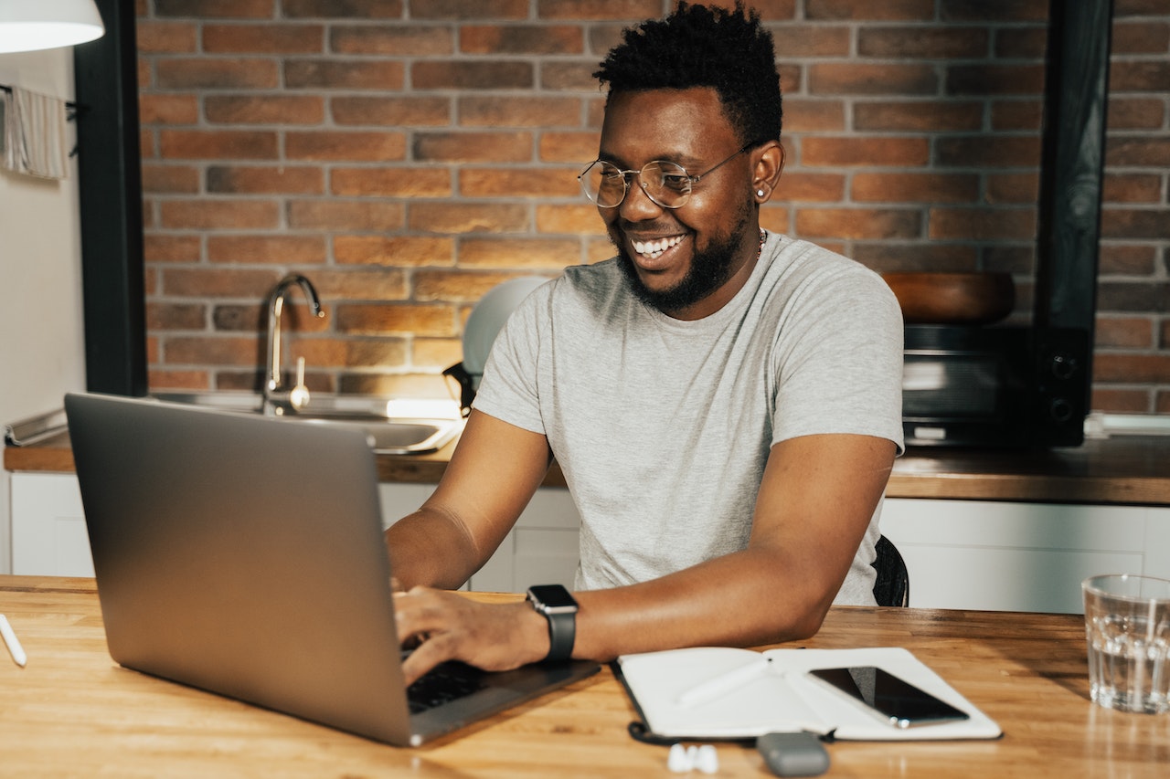 Black man is laughing while working on the laptop in the kitchen.