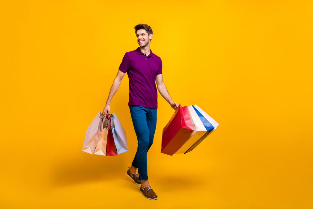 Full length body size view of attractive cheerful content guy in purple t-shirt carrying new things in colorful packages