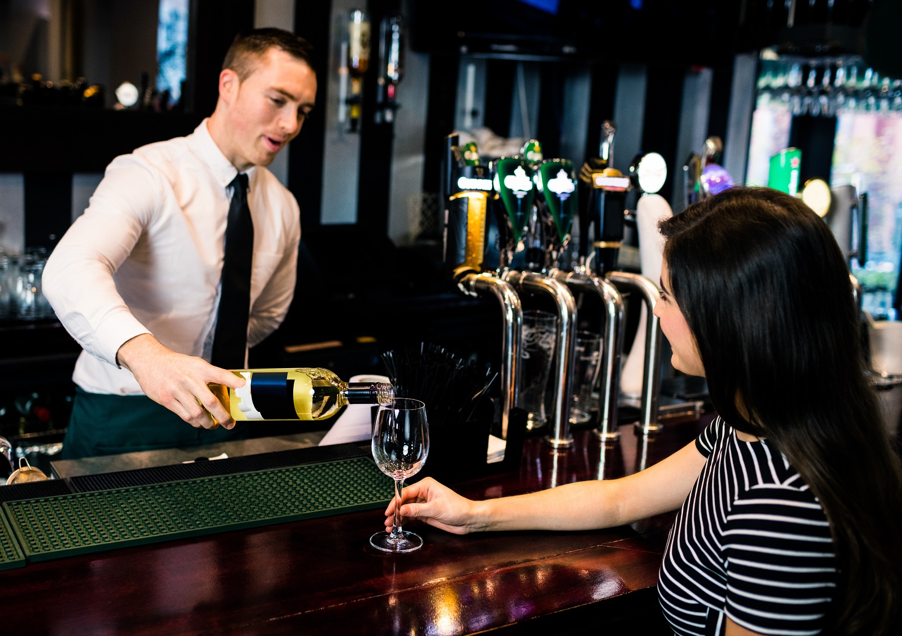 Waiter is serving a glass of wine to a woman holding a wine glass in hand.