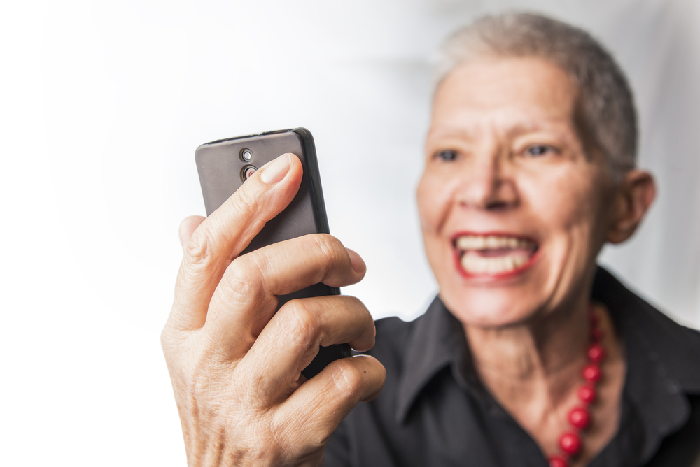 Angry senior old woman yelling, showing her cell phone, furious with customer service, in gray shirt and pearls