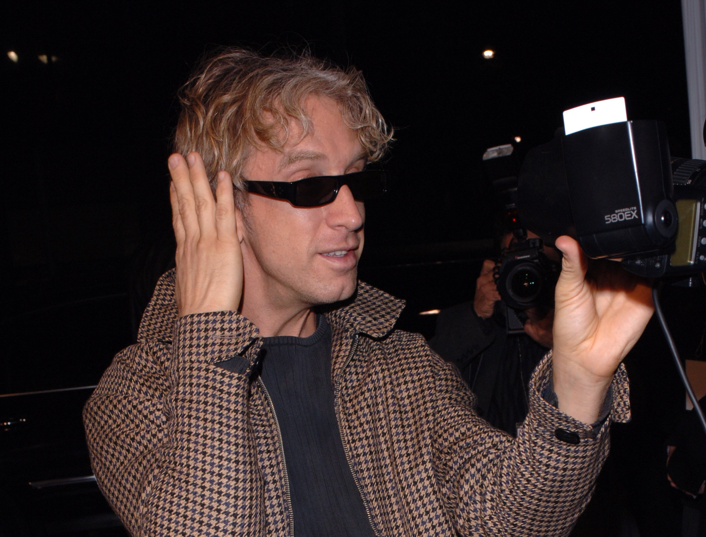 Andy Dick wearing brown jacket and glasses is talking in front of a camera - 2005