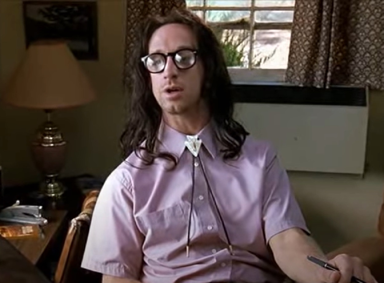 Screenshot: Andy Dick with long hair is seating on the desk - from Road Trip (2000)
