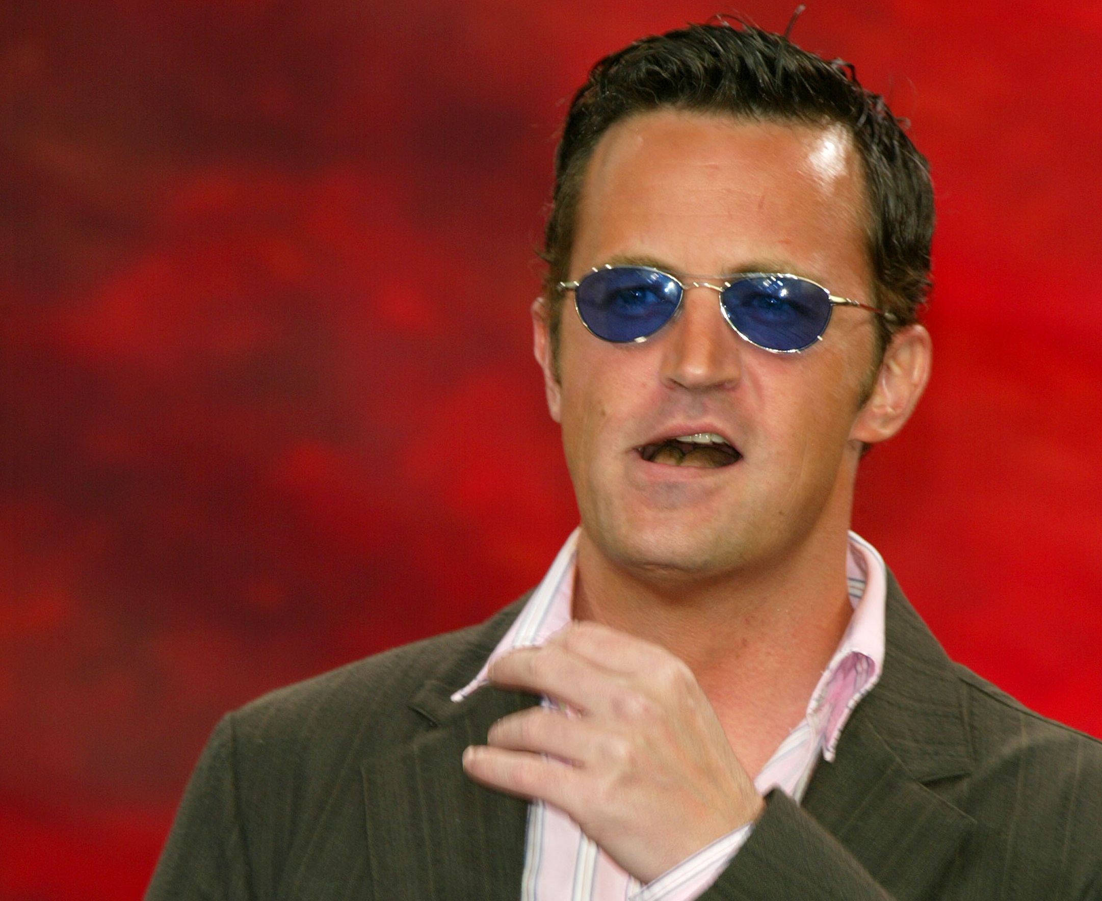 Matthew Perry wearing white shirt, green jacket and blue sunglasses is looking at side happy on red background. - 2004
