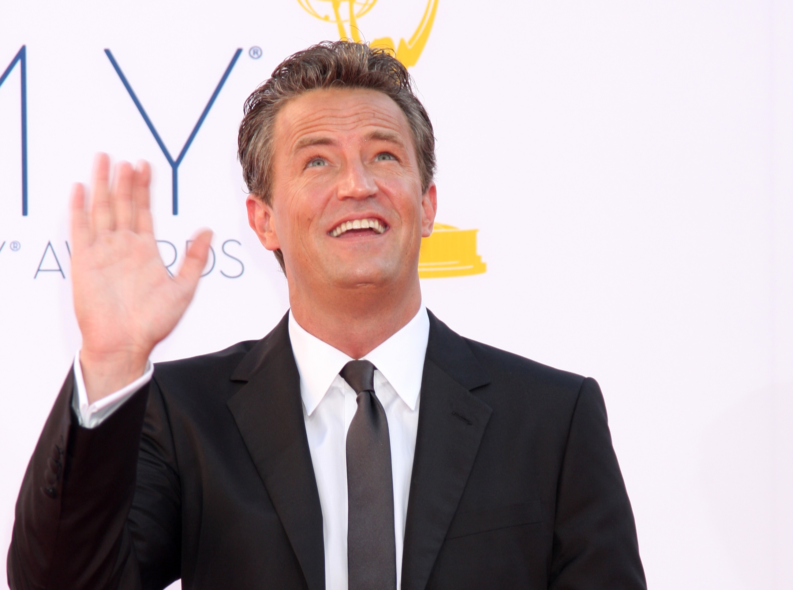 Matthew Perry wearing black suit and tie is smiling and looking up, waving with hand - 2012