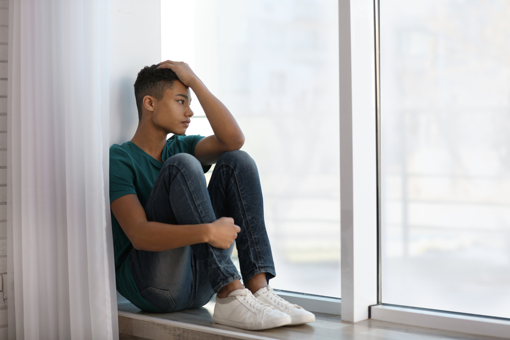 Upset African-American teenage boy sitting alone near window in green t-shirt