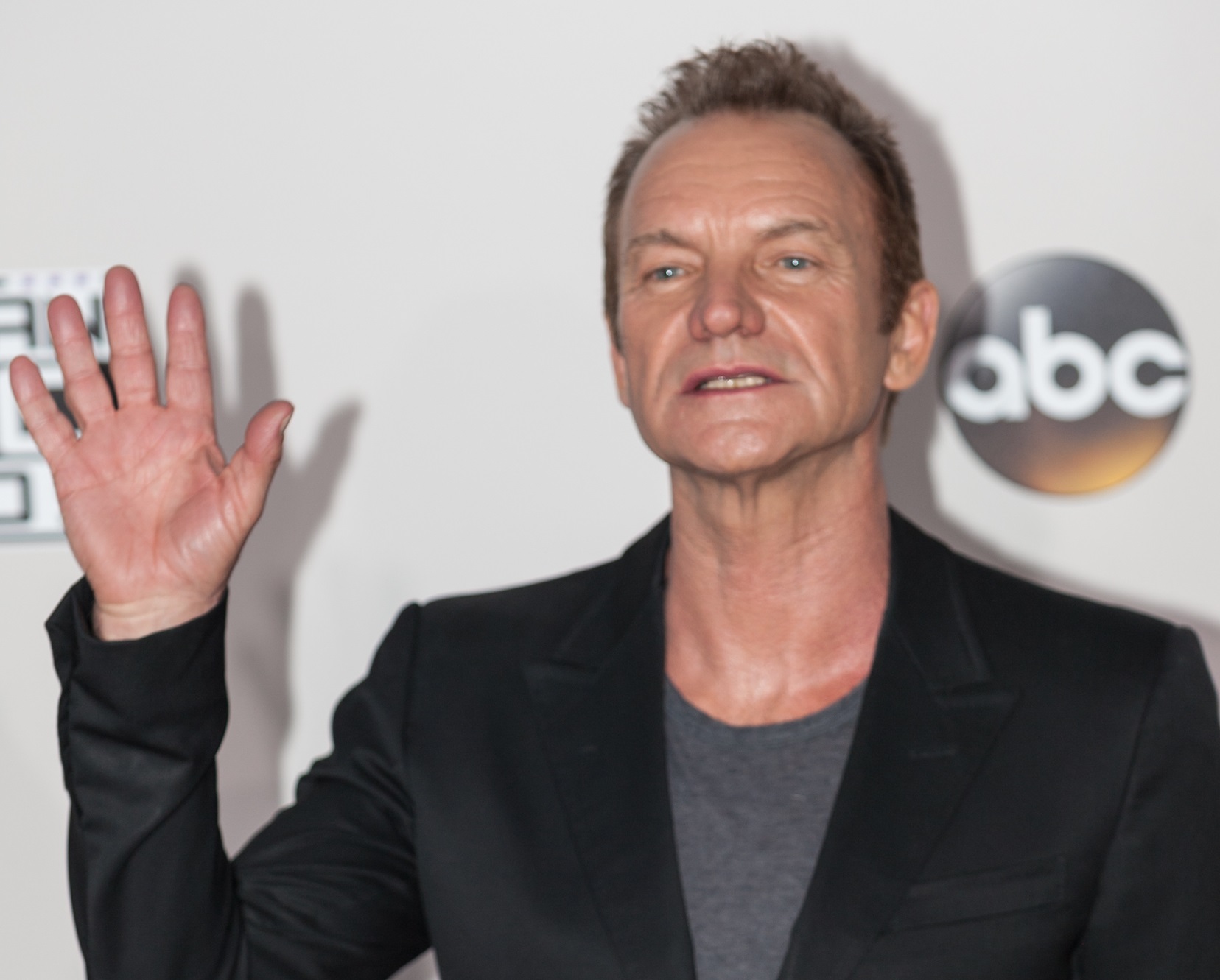 Sting wearing grey shirt and black jacket smiling and making a hand gesture to camera - 2016