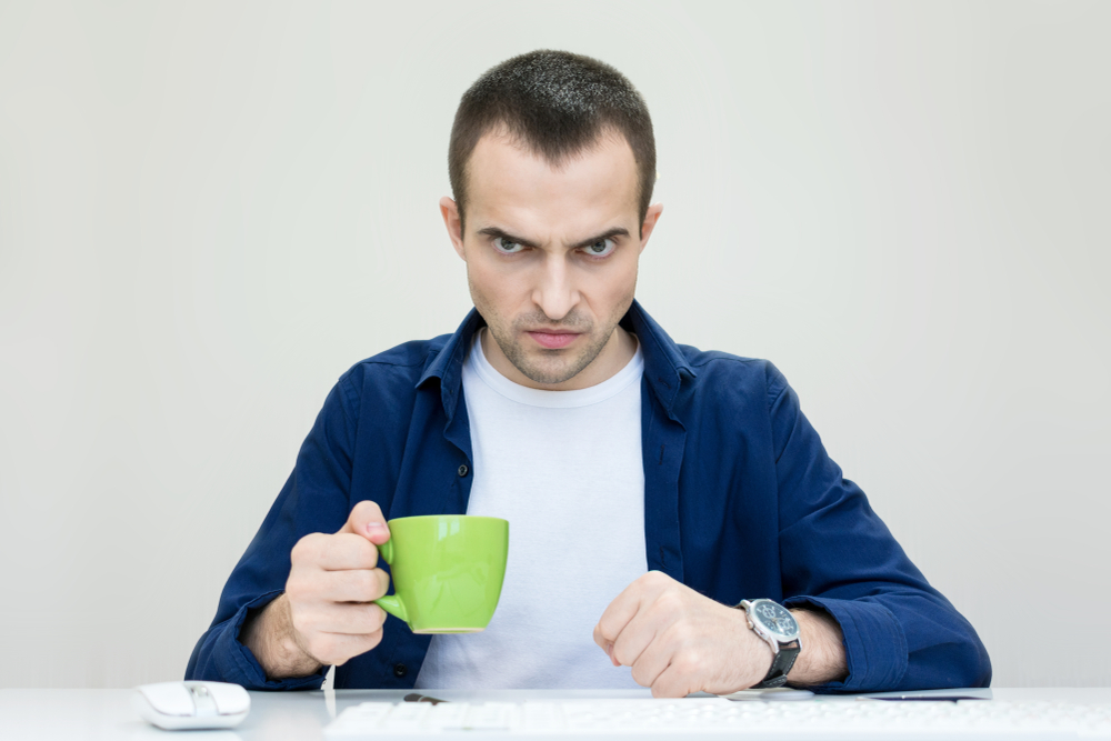 Angry man in blue shirt holding a cup of coffee looking at the computer