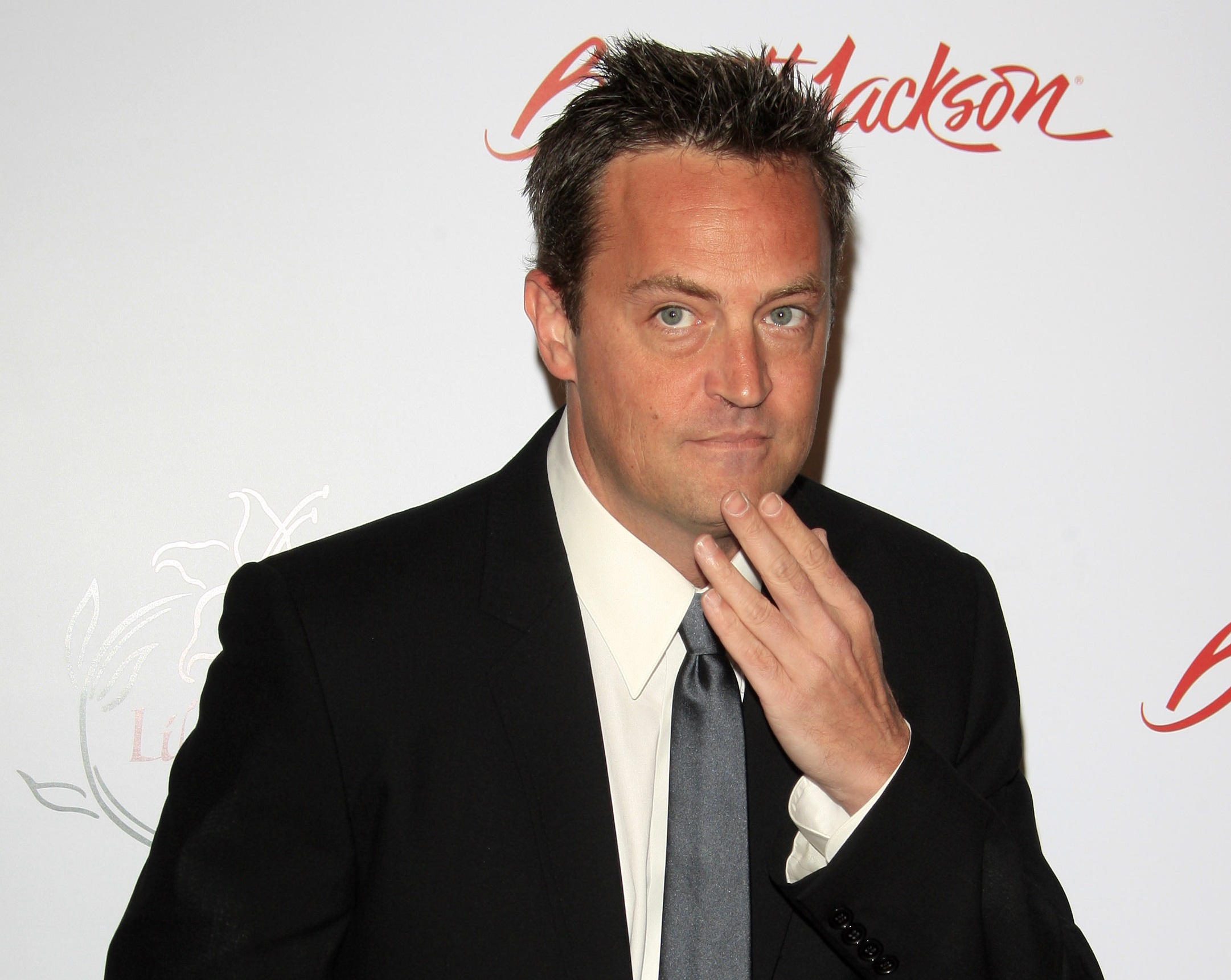 Matthew Perry wearing black suit and grey tie is touching his chin and looking upset - 2008