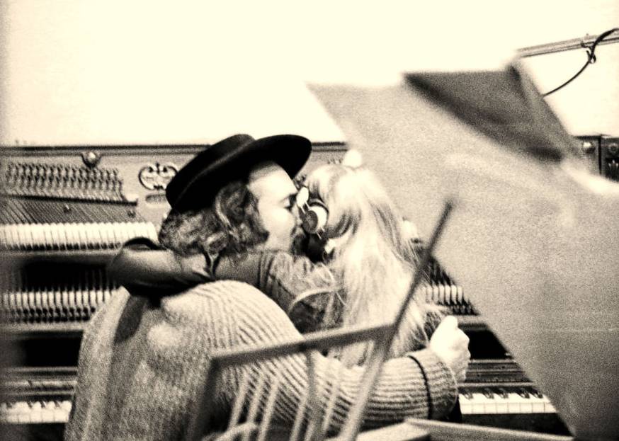 David Crosby holding Joni Mitchell while both are seated at an upright piano