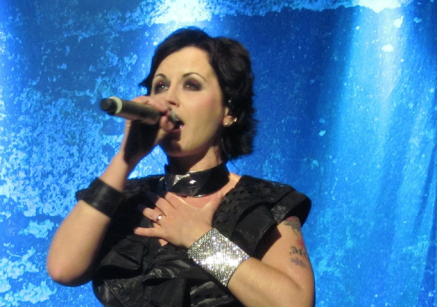 Dolores O'Riordan wearing black dress and silver bracelet is performing on stage on blue background - 2012