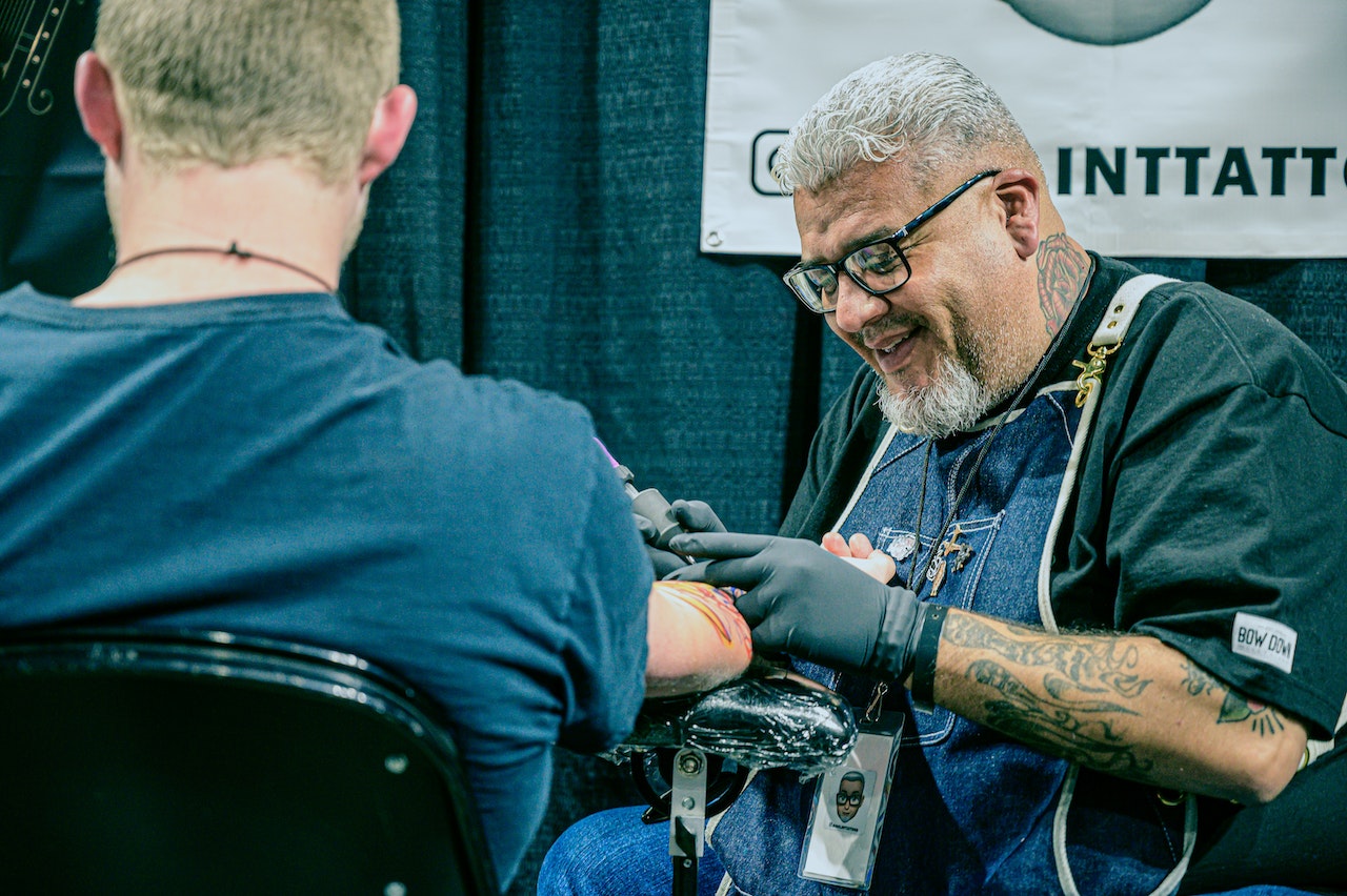 Man in a blue shirt is getting a new tattoo from tattoo artist.