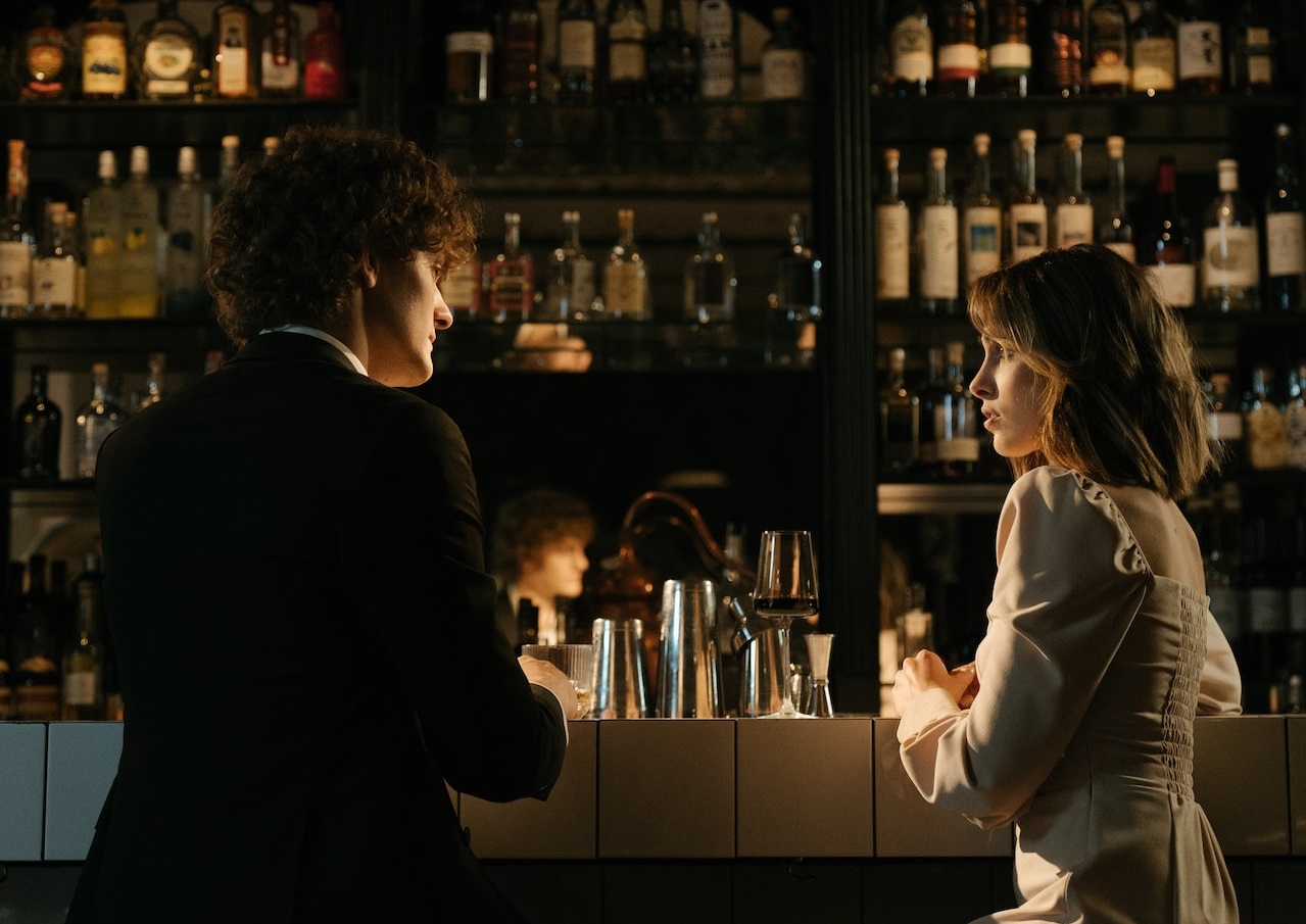 Man in black suit is talking with a woman seating in the bar.