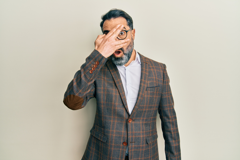 Middle age teacher  with beard and grey hair wearing business jacket and glasses peeking in shock covering his face and eyes with hand, looking through fingers
