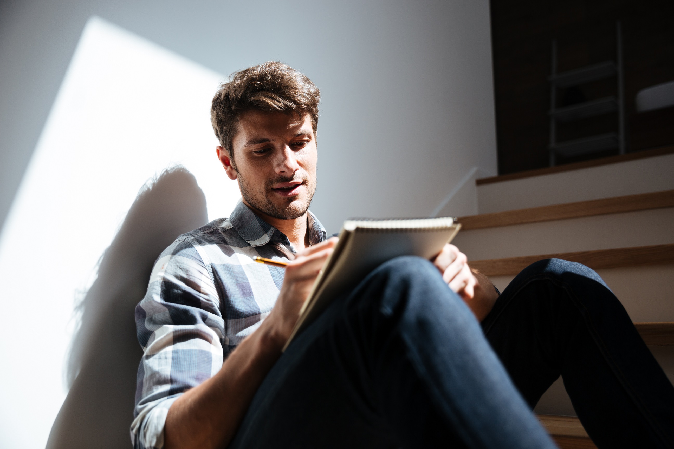 Man in checkered shirt and jeans sitting on stairs at home and writing a letter.