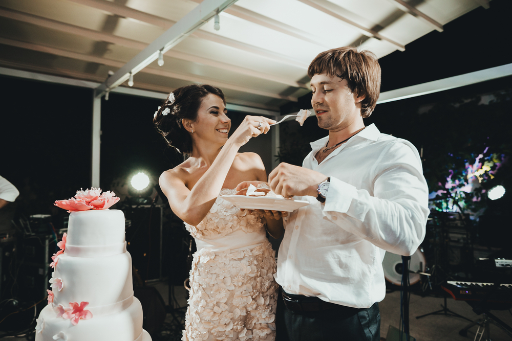 Bride holds fork with wedding cake in front of  groom's face