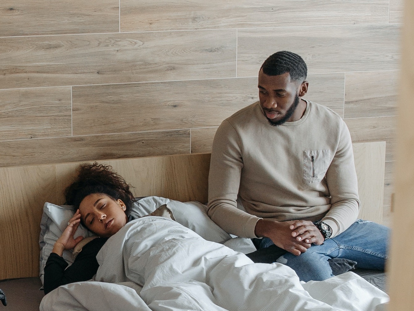 Man is seating in bed next to a sick woman who is sleeping on the bed.
