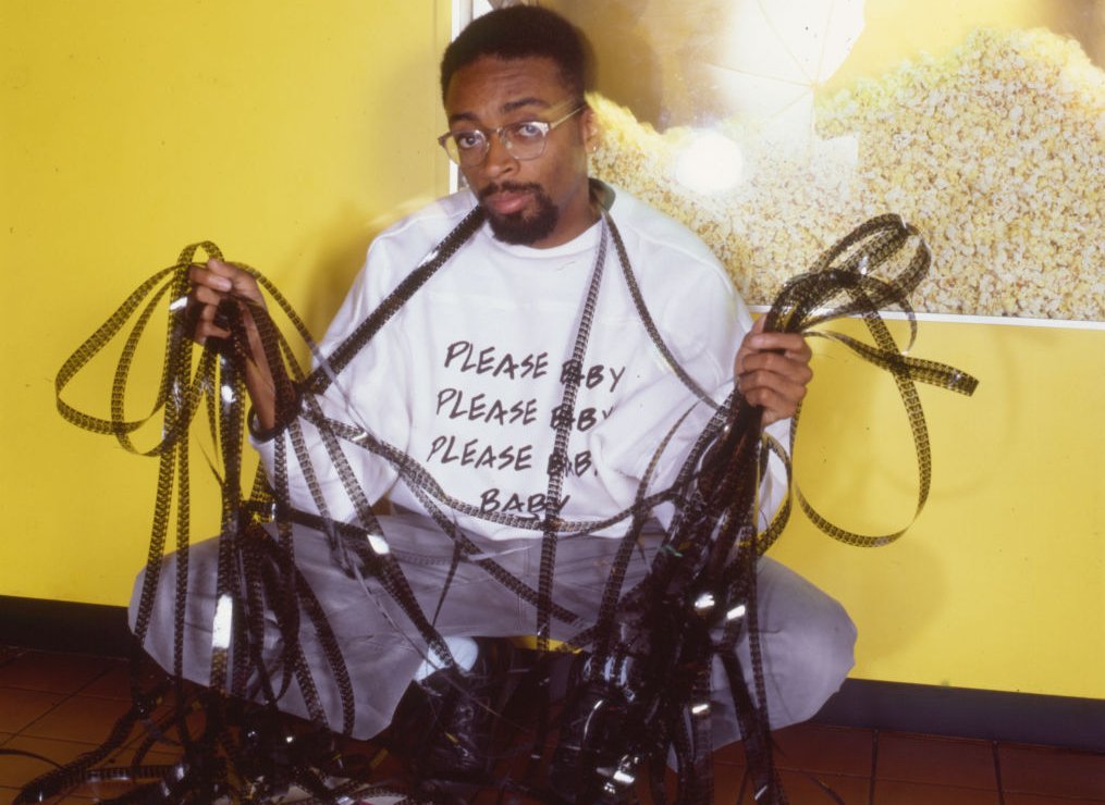 Portrait of African American film director Spike Lee, holding film reels, while crouching in front of a poster for his film 'She's Gotta Have It', New York, 1986