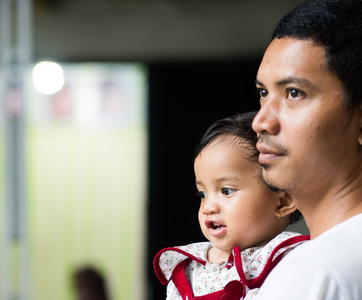 Father wearing white shirt holding his baby wearing red dress and looking a side,