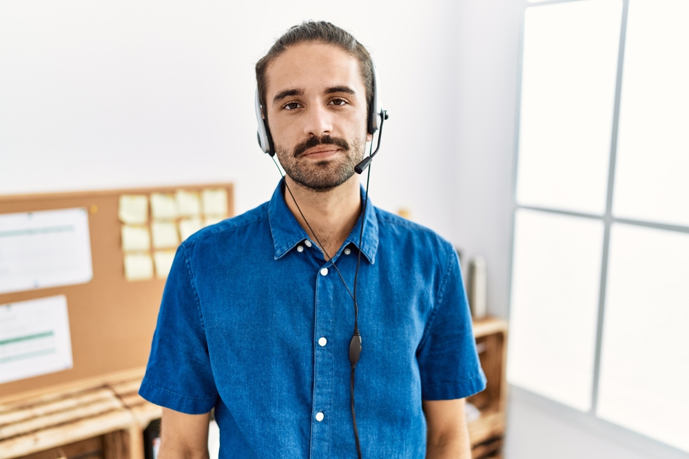 Young Hispanic man with beard wearing call center agent headset at the office with serious expression on face