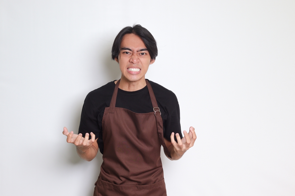 Portrait of annoyed Asian barista man in brown apron making angry hand gesture with fingers on gray background