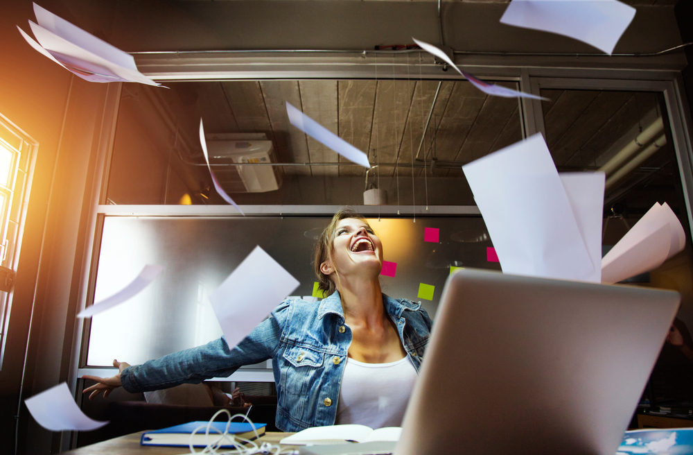 Happy woman sitting on a desk throwing papers in air
