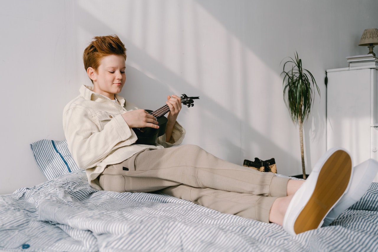 A boy playing at ukulele on the bed.