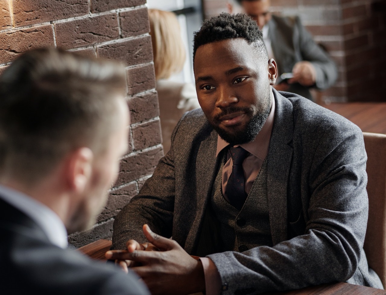 Two man wearing suits talking to each other and seating on the table.