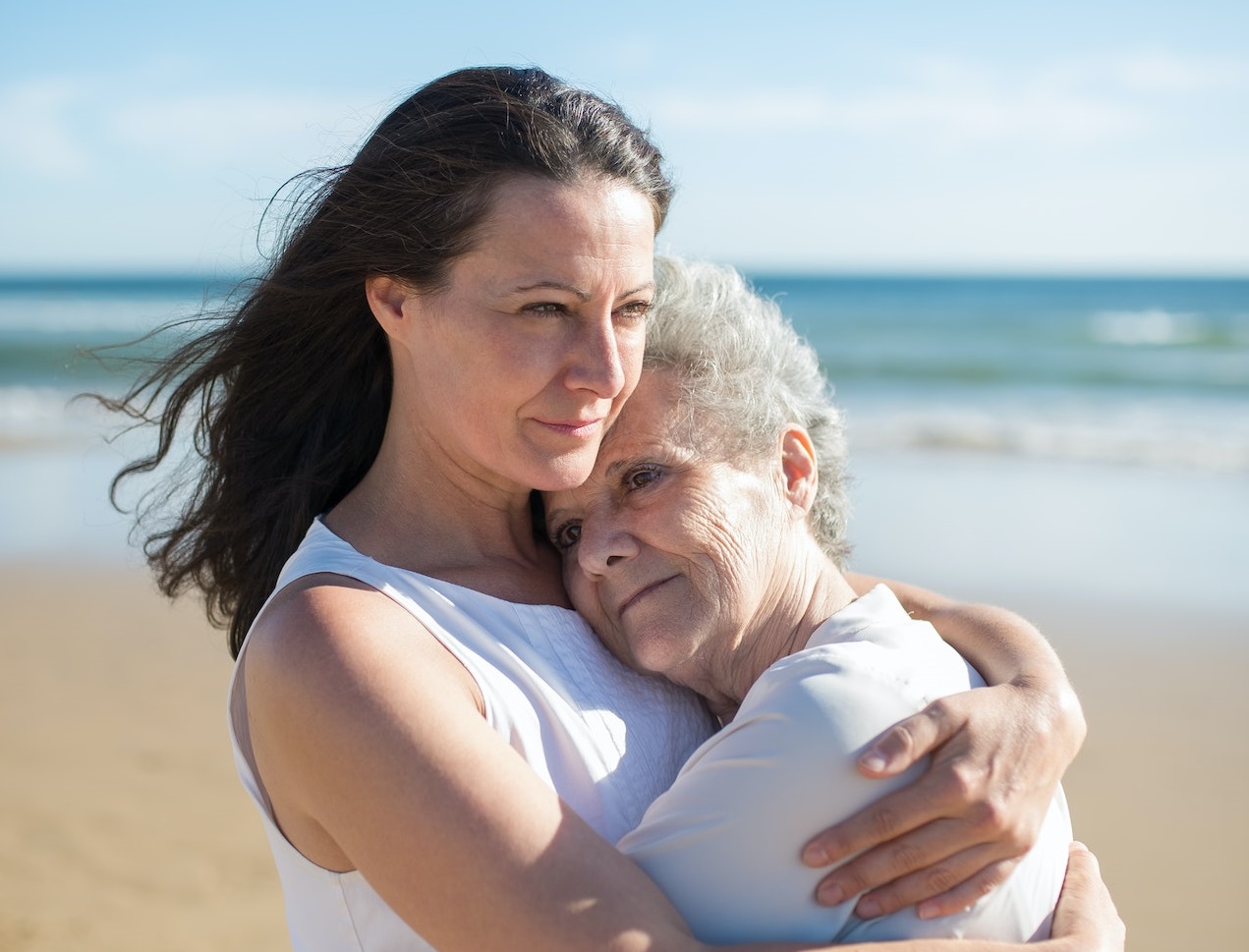 Younger woman and elder woman wearing white dresses hugging each other on the beach.