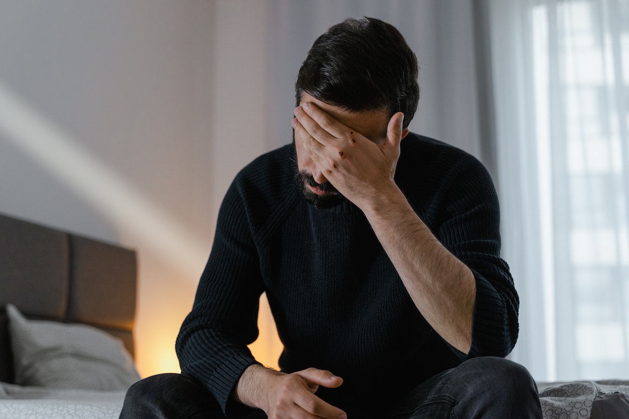 Man wearing black shirt seating on the bed and covering his face with hand.