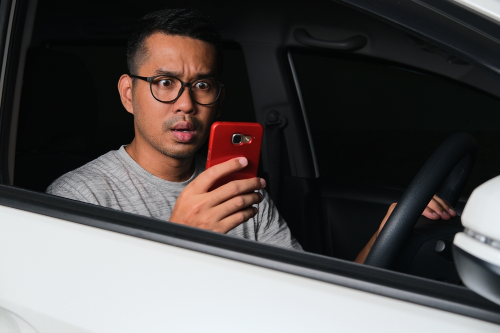 Asian man looking to his phone with shocked expression when driving a car