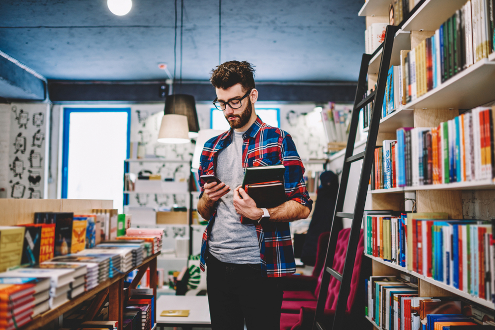 Handsome hipster guy in spectacles making money transaction for buying new bestseller via mobile phone