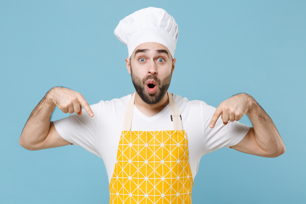 Shocked chef in hat and apron pointing at himself over blue background