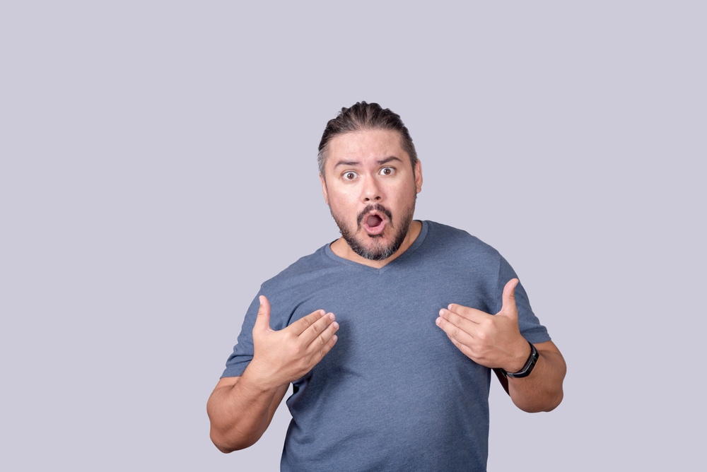 A man disagrees with the accusations thrown at him, in gray t-shirt over gray background looking shocked