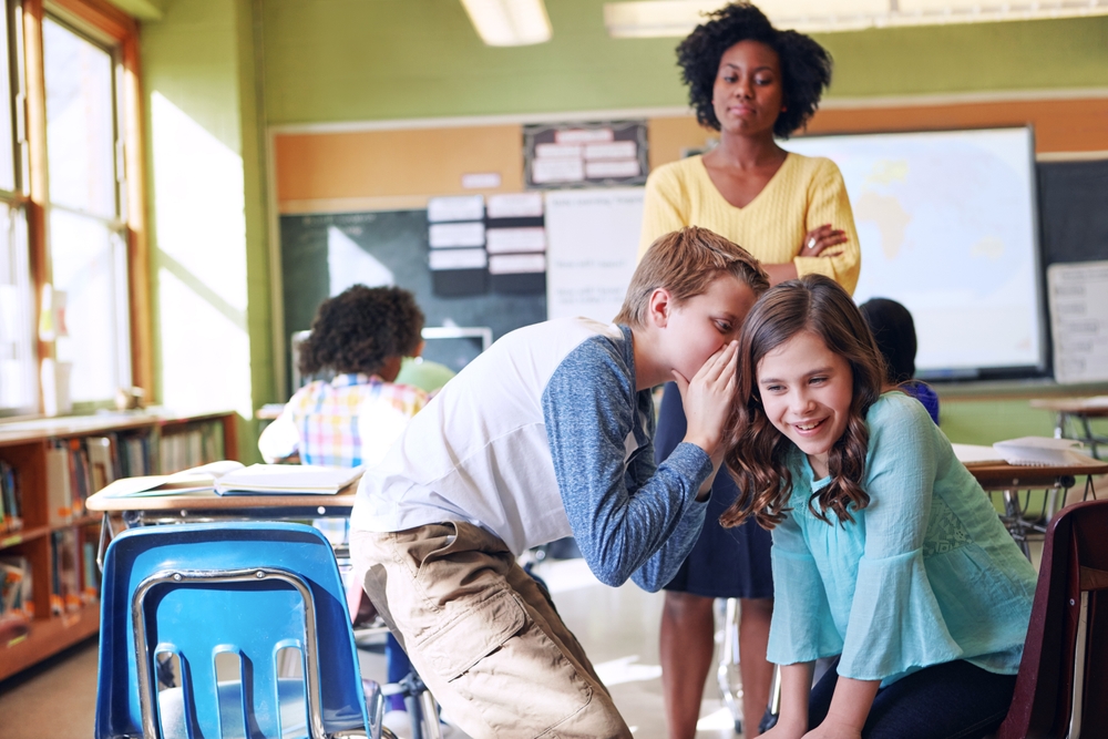 Students  whisper in classroom with teacher standing in front of them ready to  discipline them