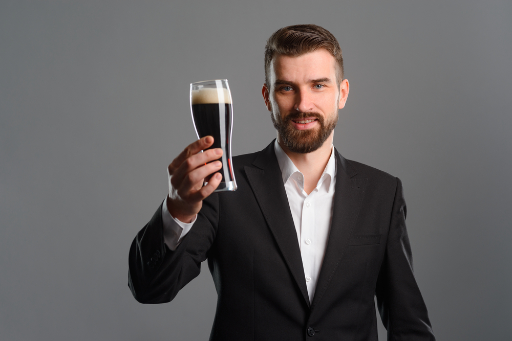 Man in suit holding a glass of drink in front of himself smiling over gray background