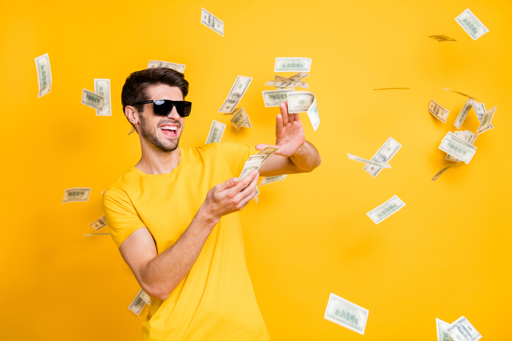 Photo of young handsome careless guy throwing  money banknotes away wearing sun specs casual t-shirt
