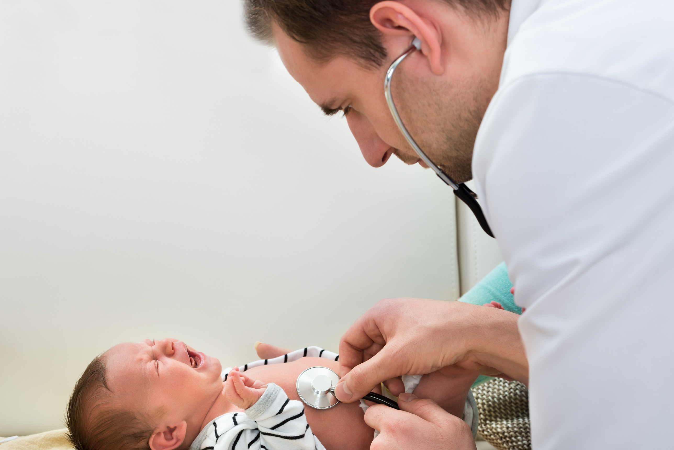 Young male doctor is examining the crying baby on hospital bed.