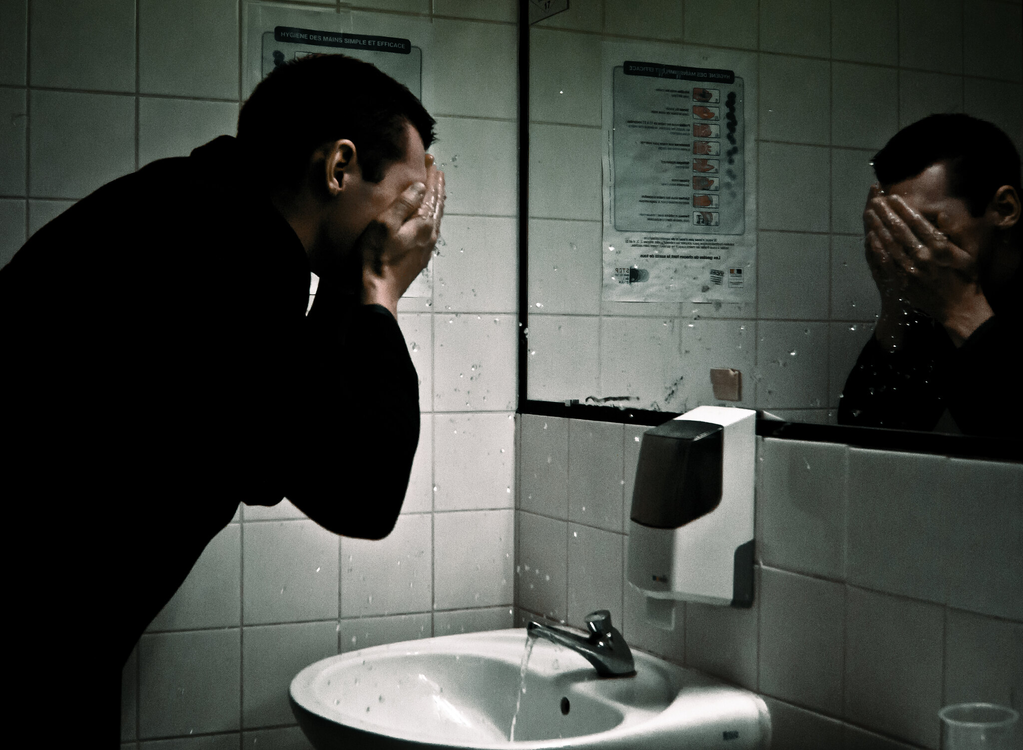 Man is washing face over sink in man's public toilet.