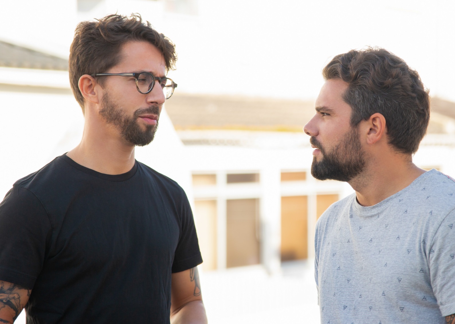Two bearded man are talking outside with upset faces.