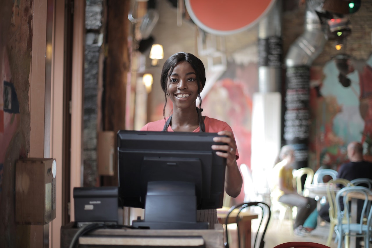 Young black woman is standing at cash register at restaurant and smiling.