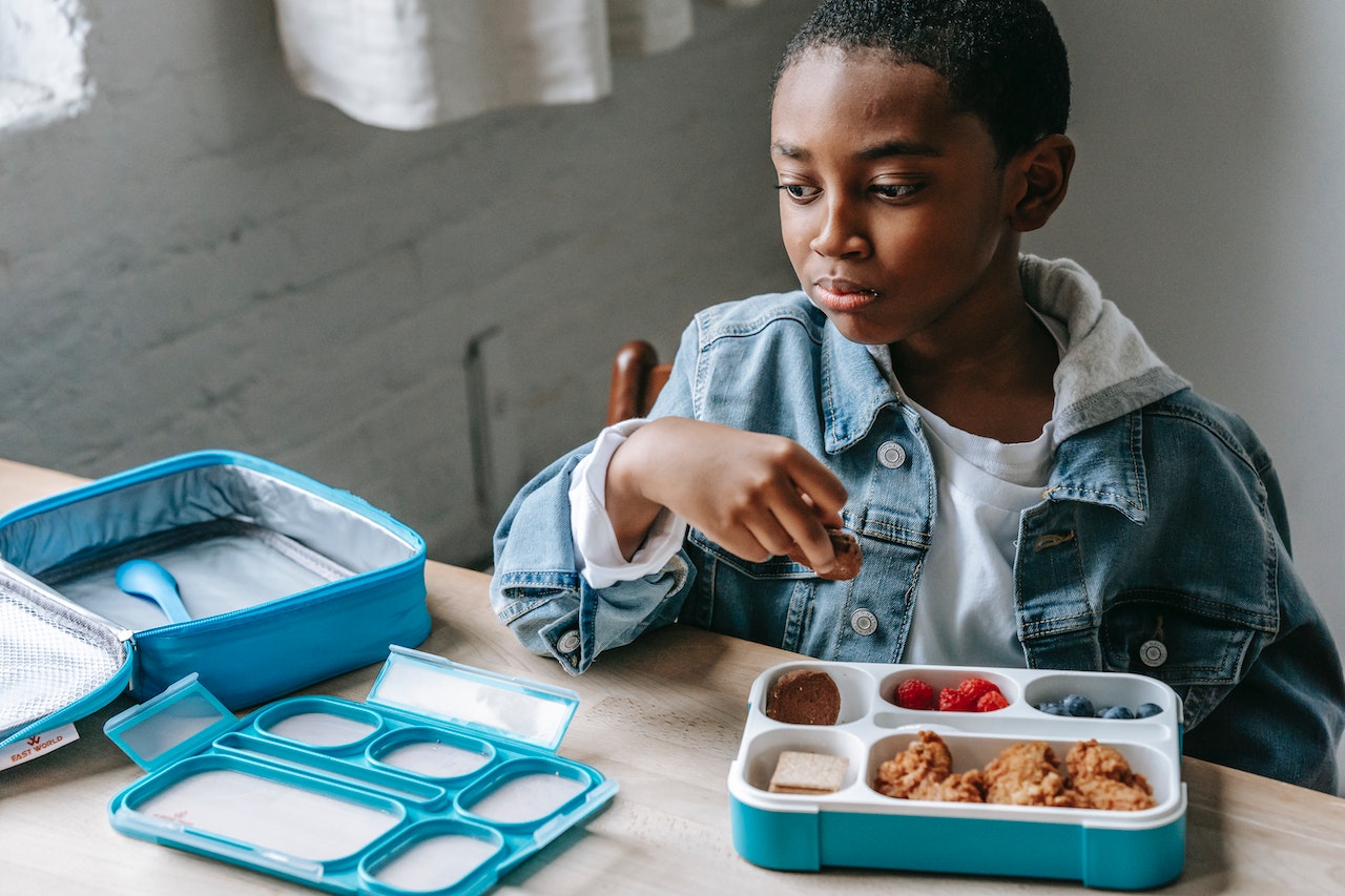 Young black kid is seating and eating lunch at school table.