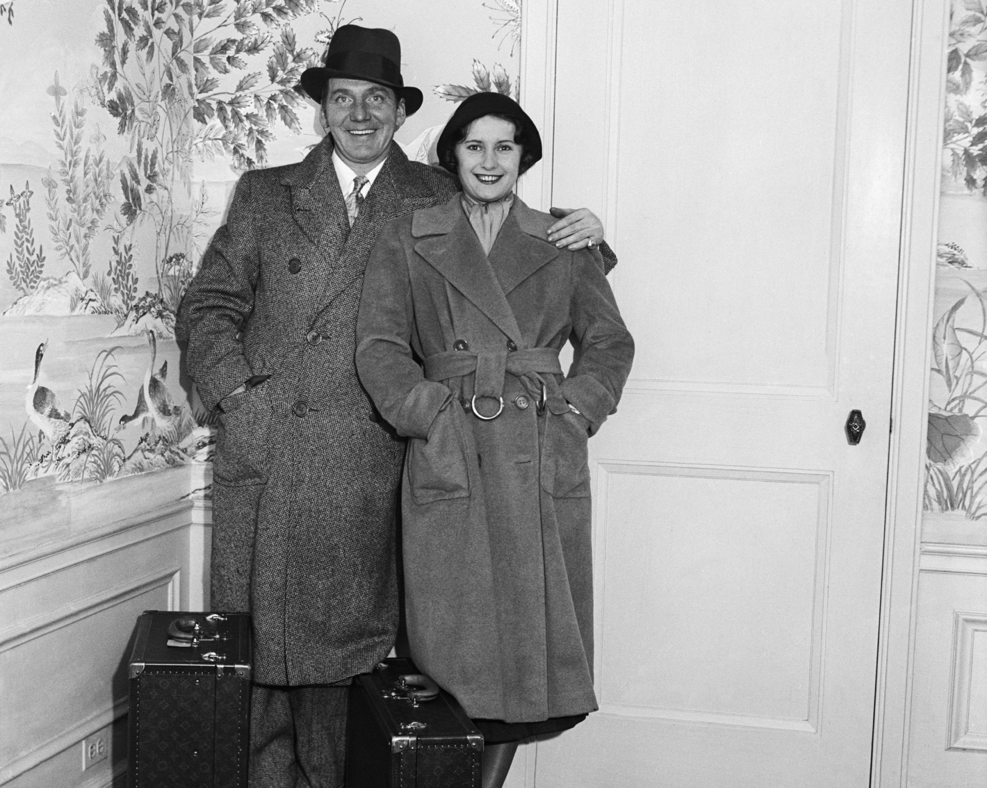 Barbara Stanwyck   and husband Frank Fay at Hotel Waldorf-Astoria in coats standing by a door
