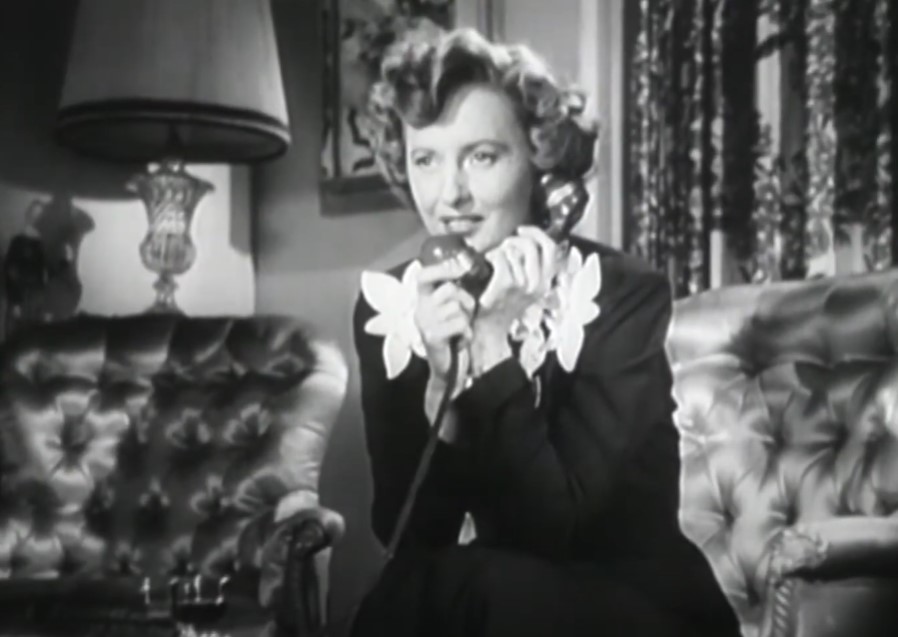Barbara Stanwyck in East Side, West Side in black dress sitting on a couch