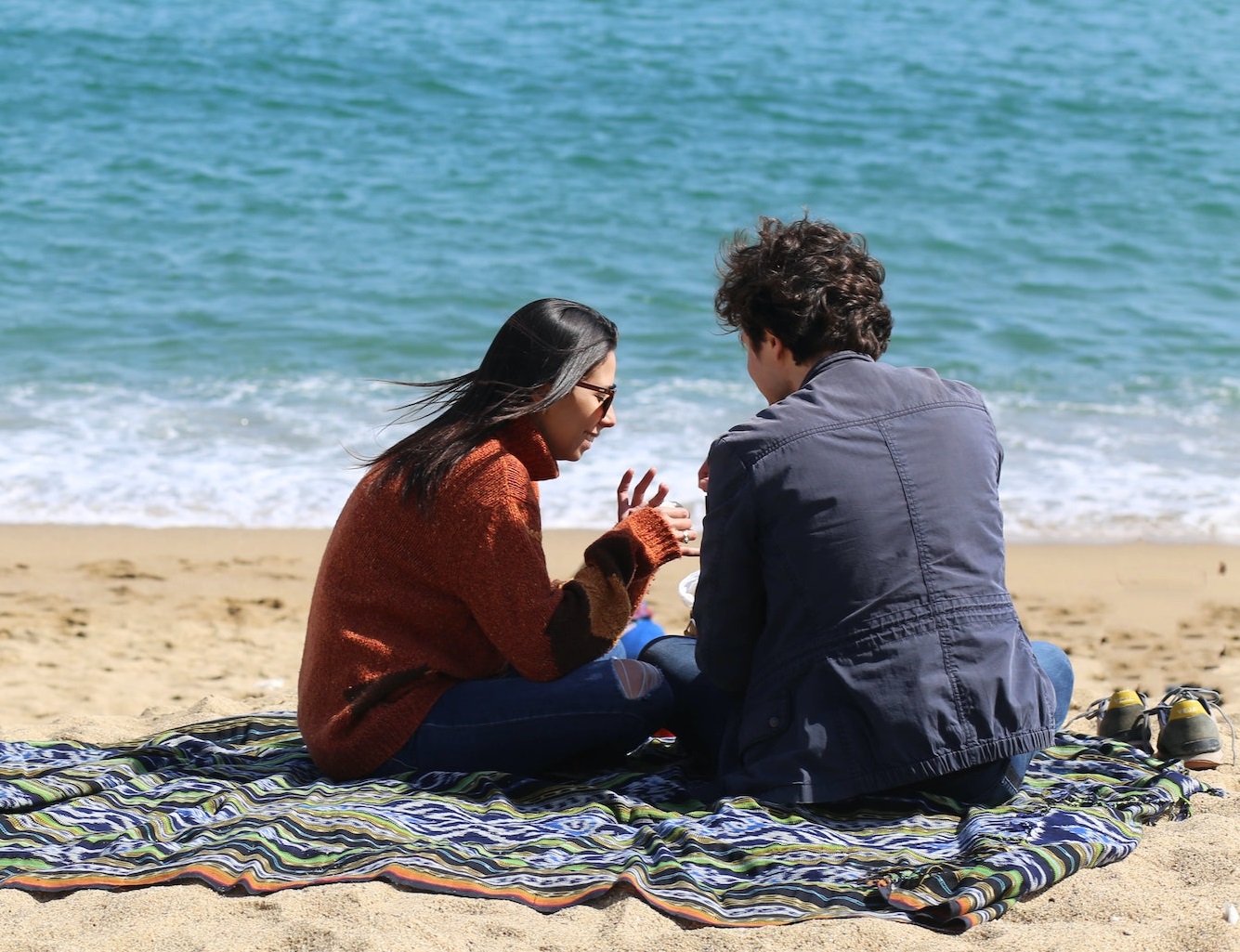 Young woman is seating on the beach next to a man wearing blue jacket.
