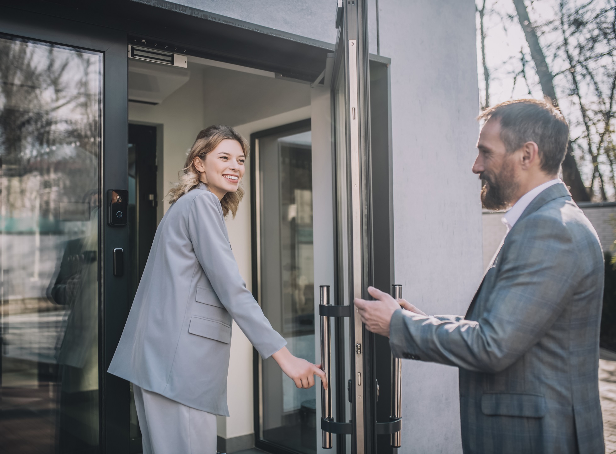 Man in grey suit is opening door for woman in grey suit to enter and smiling.