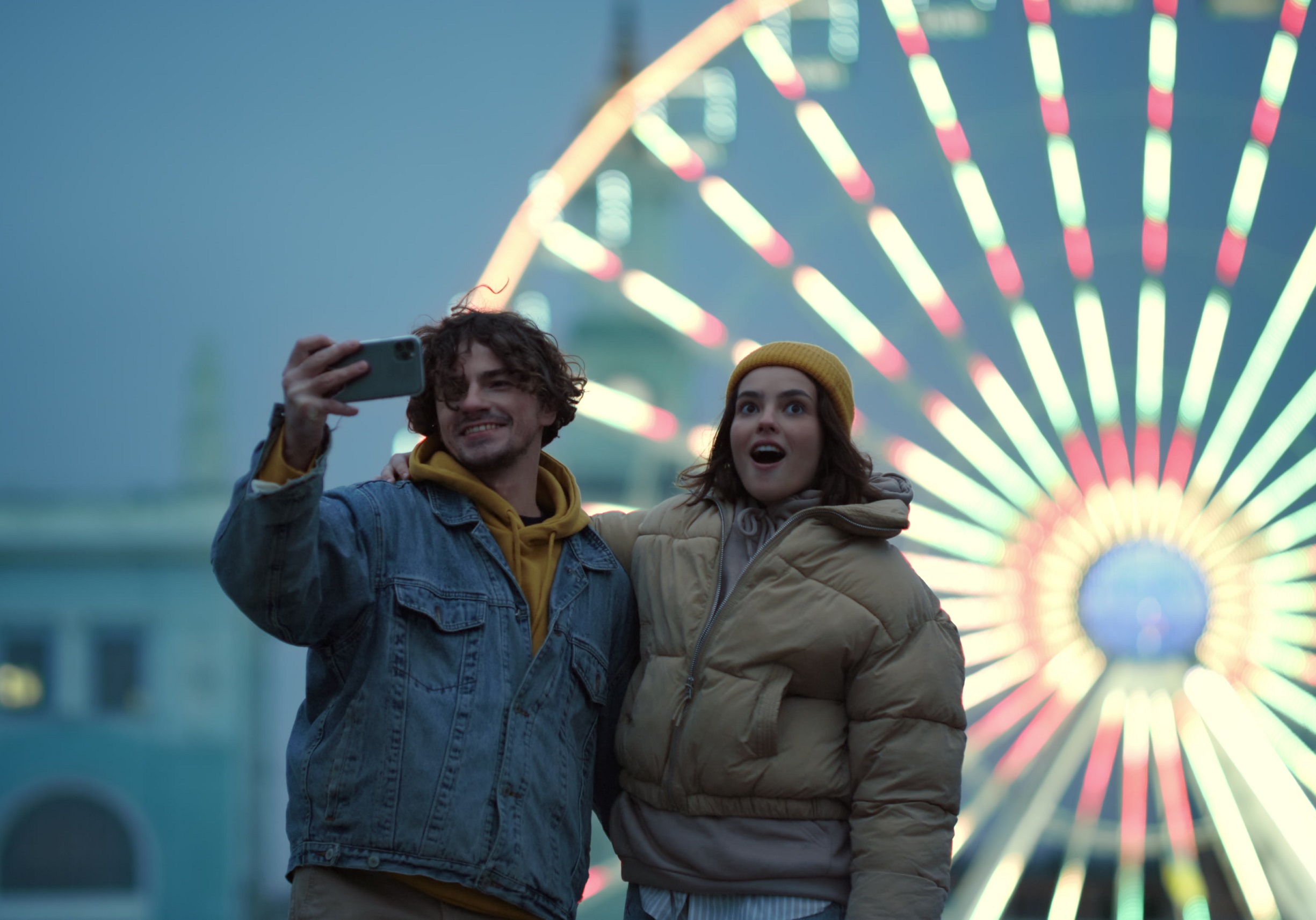 Couple is taking a selfie on cellphone against illuminated Ferris wheel.