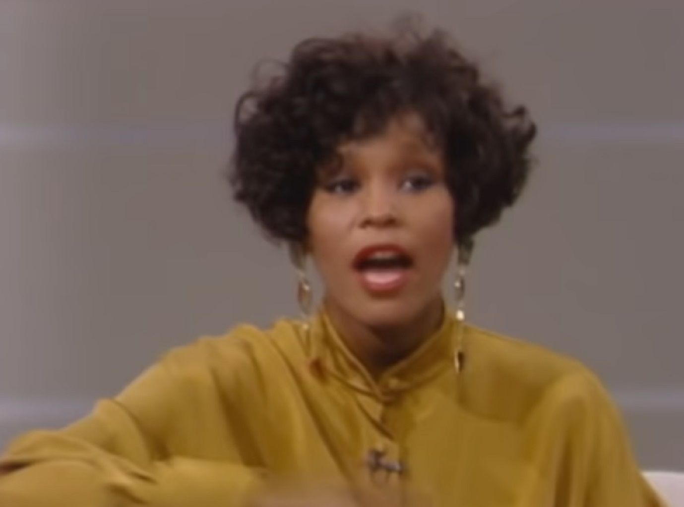Screenshot of Whitney Houston wearing orange shirt is talking - from Whitney (2018).